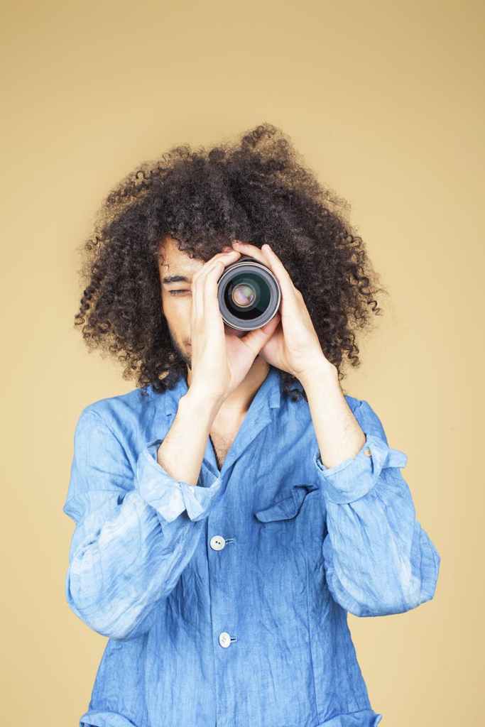 Person with curly hair holding a camera lens towards their face, wearing a blue shirt, against a beige background.