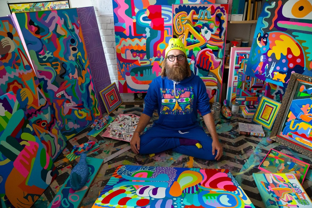 An artist sitting cross-legged in a vibrant studio filled with colorful abstract paintings and artworks.