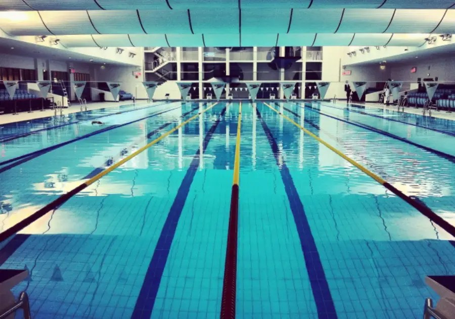 Indoor swimming pool with clear water, marked lanes, and diving blocks. The area is bright with natural light coming through large windows.