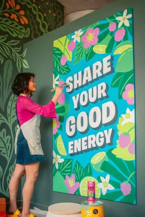 A woman painting a vibrant mural that reads 'SHARE YOUR GOOD ENERGY' surrounded by colorful flowers and leaves.