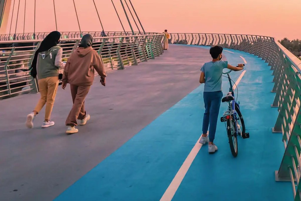 A boy walking his bicycle on a blue pathway alongside two women jogging in the evening light, with lush greenery in the background.
